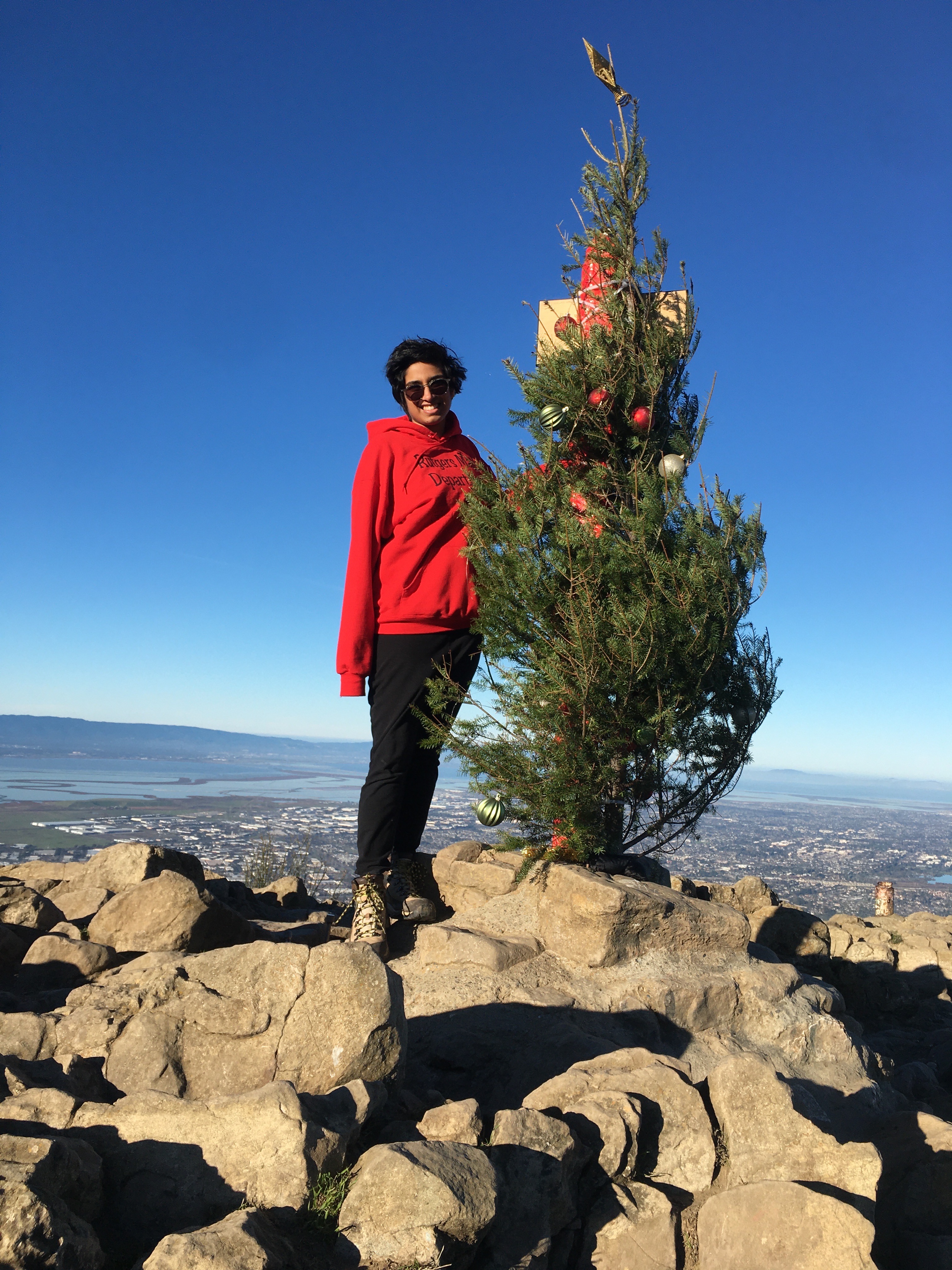 Karuna wearing a Rutgers Math sweatshirt while standing next to a Christmas tree at the top of Mission Peak in Fremont, CA.